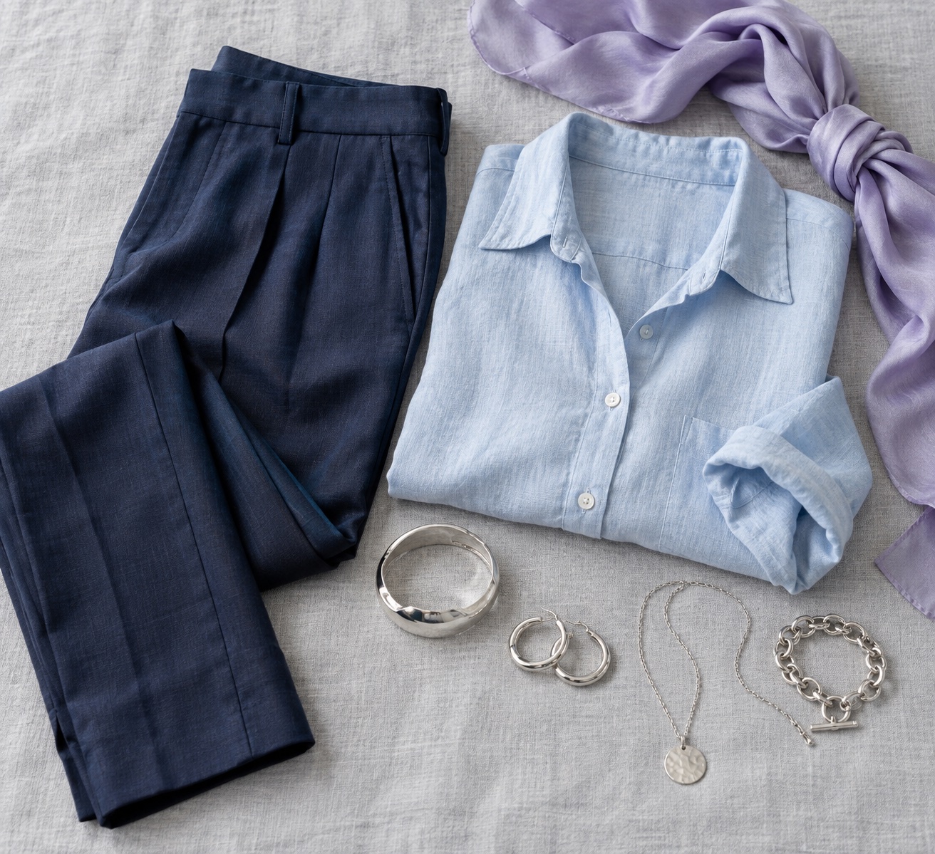 Flatlay of soft navy trousers, powder-blue linen shirt, lavender silk scarf, and silver jewelry on pale gray fabric